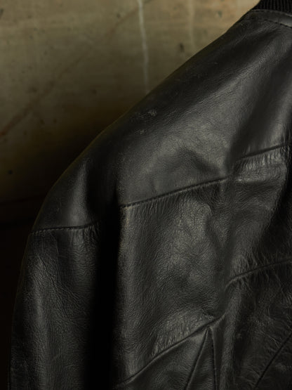 Close-up of a black leather jacket showing the texture and stitching on the shoulder and upper sleeve, set against a dark concrete background.