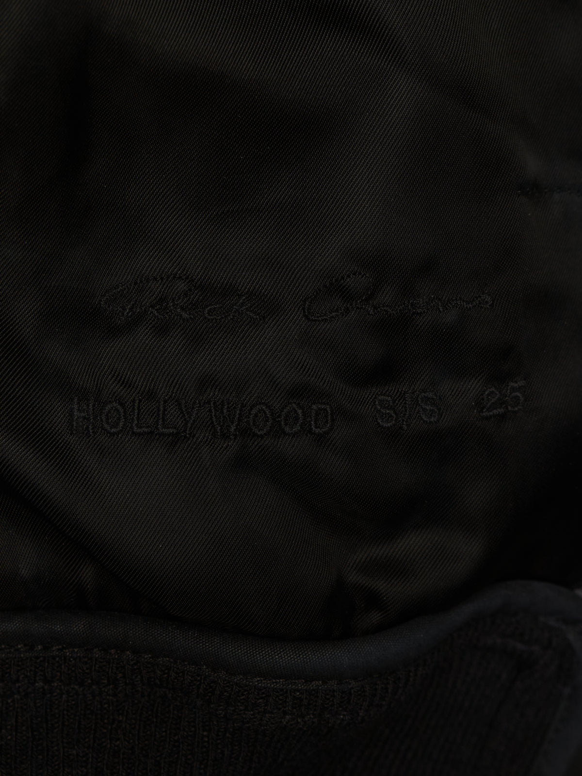 Close-up of a black fabric surface with faint embroidered text reading 'Rick Owens' and 'HOLLYWOOD S/S 25' on a dark satin-like material.