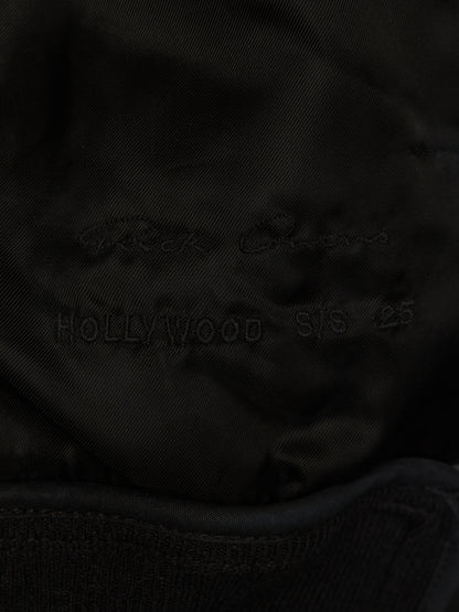 Close-up of a black fabric surface with faint embroidered text reading 'Rick Owens' and 'HOLLYWOOD S/S 25' on a dark satin-like material.