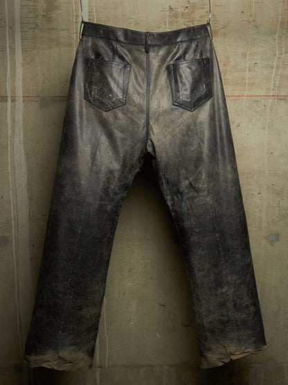 Distressed black leather pants with a faded, weathered finish, featuring two back patch pockets, shown hanging against a textured concrete wall.