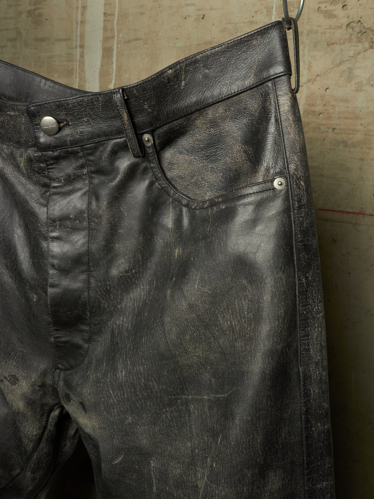 Distressed black leather pants hanging against a textured concrete wall, showing a heavily worn, vintage finish with scuffed details and silver-toned rivets.