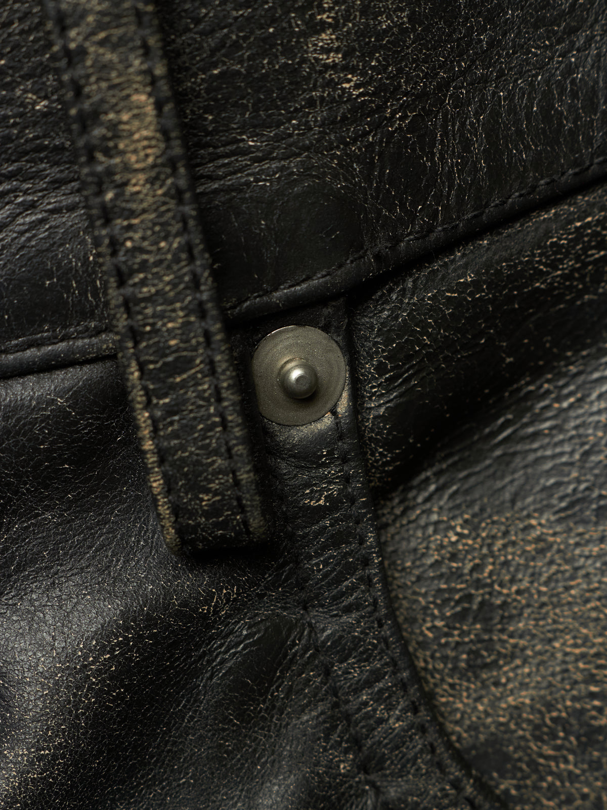 Close-up of a distressed black leather garment, likely a jacket or vest, featuring a metal snap button closure and visible gold-toned wear effects.
