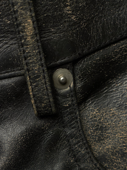 Close-up of a distressed black leather garment, likely a jacket or vest, featuring a metal snap button closure and visible gold-toned wear effects.