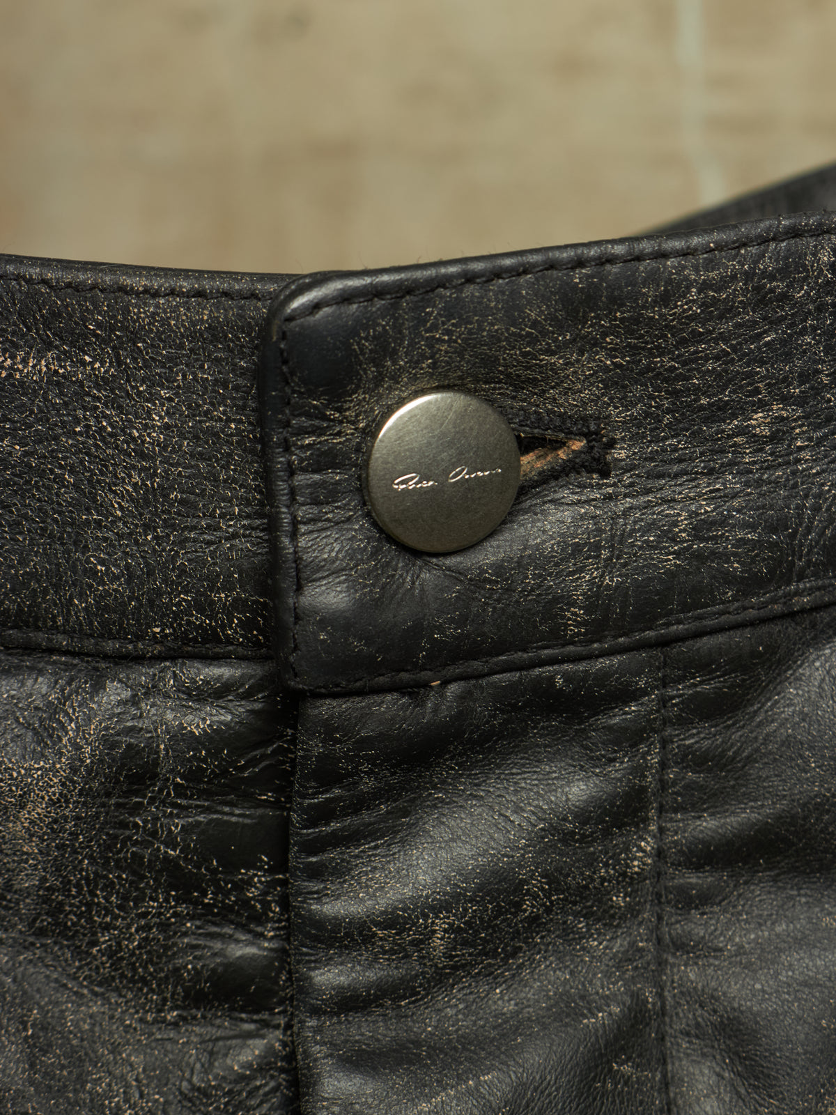 Close-up of a distressed black leather garment, likely pants or shorts, showing a round metal button with the brand name 'Gian Lorenzo' engraved on it.