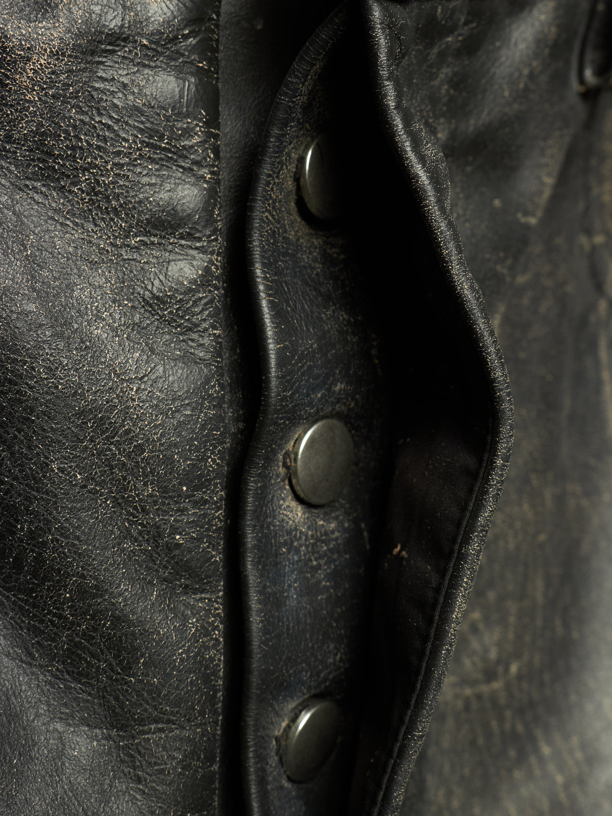 Close-up detail of a black leather jacket showing the texture of the leather, a buttoned flap, and dark metal snap buttons.
