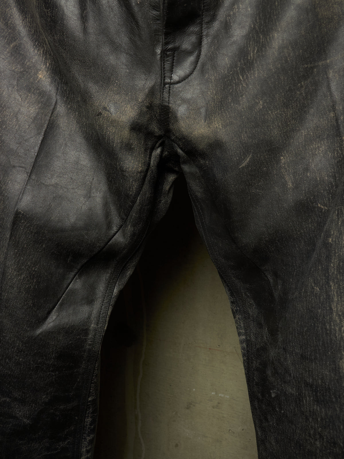 Close-up of black leather pants showing the front fly area, seams, and a light-colored floor beneath.