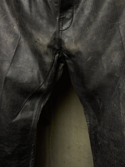 Close-up of black leather pants showing the front fly area, seams, and a light-colored floor beneath.