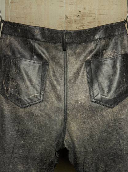 Distressed black leather shorts with a distressed finish, featuring two back patch pockets and a central seam, shown hanging against a neutral background.