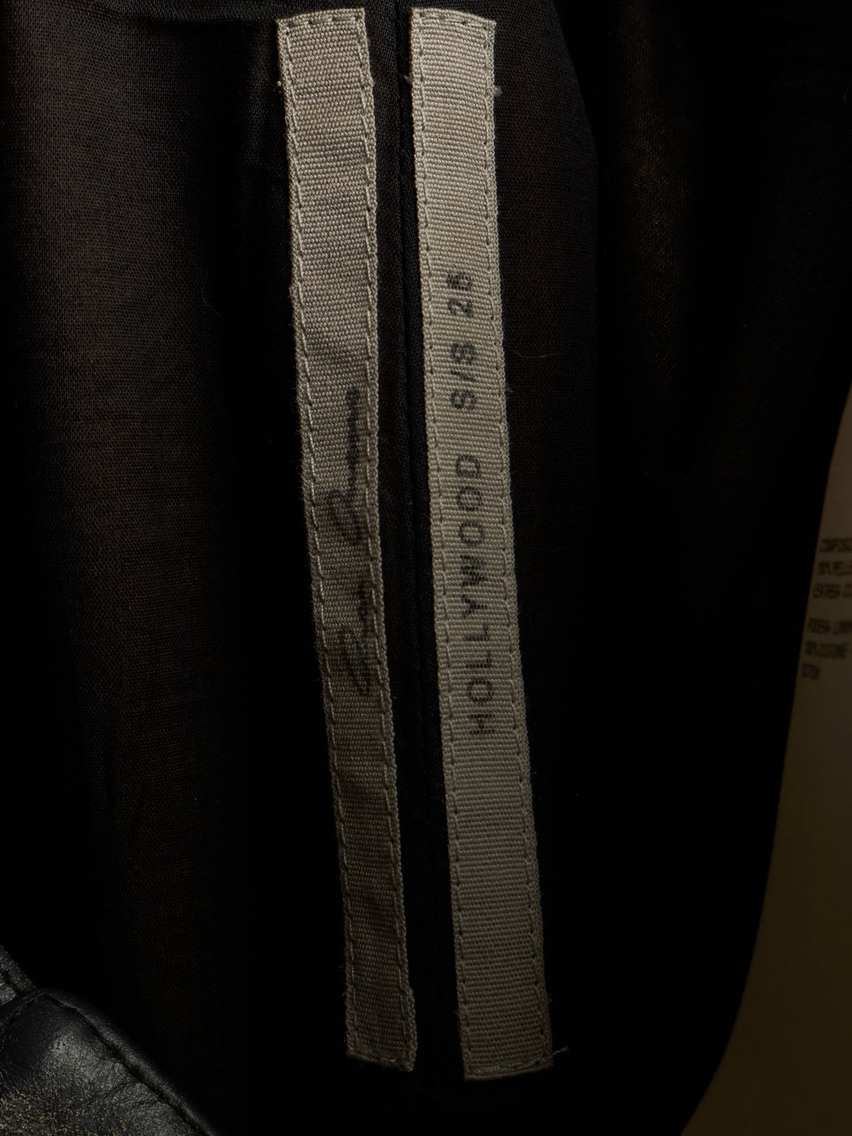 Close-up of a black garment's interior label area showing two vertical gray fabric strips with printed text including 'HOLLYWOOD S/S 26'.