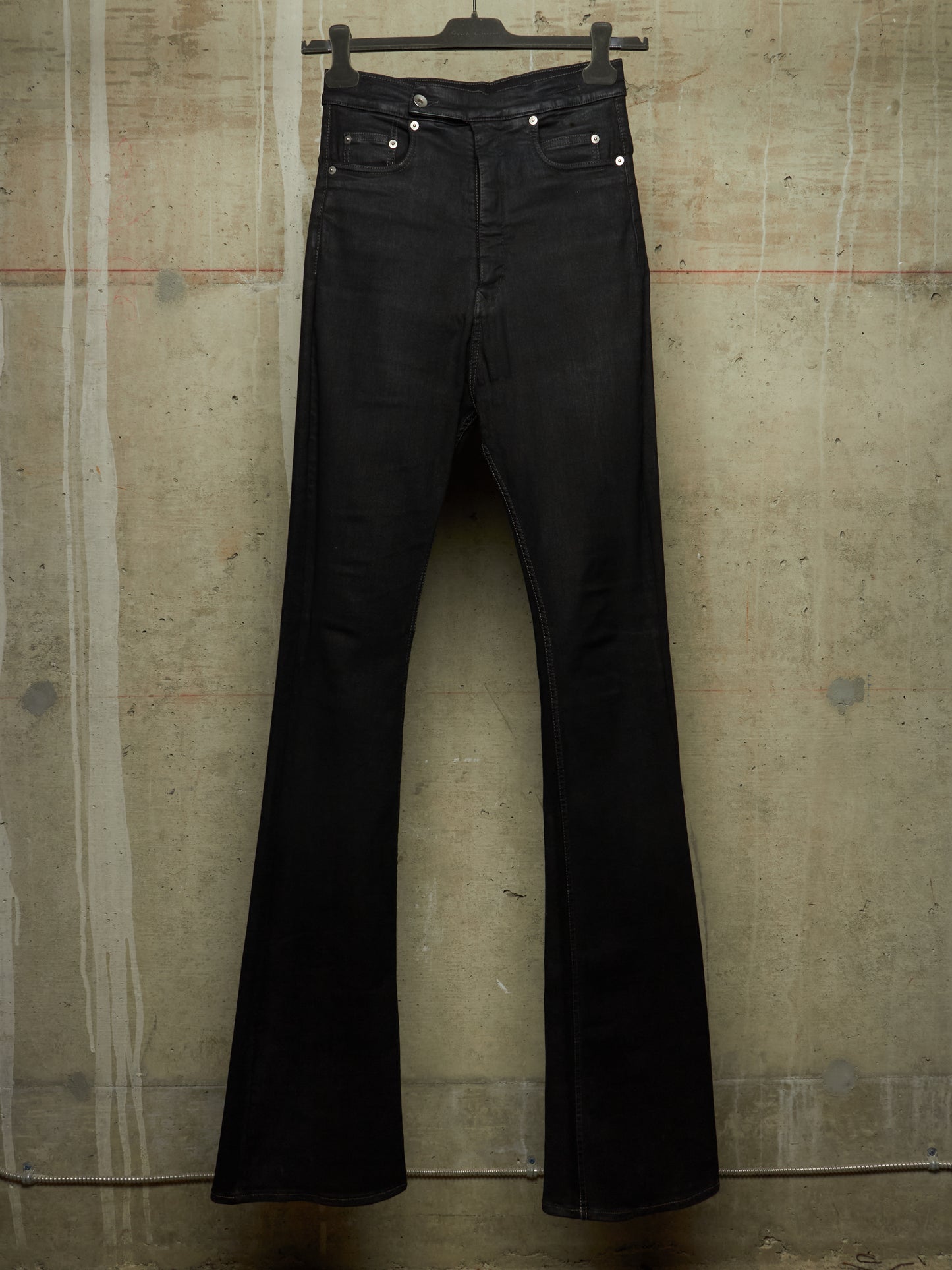 Black flare jeans hanging on a black hanger against a textured concrete wall.
