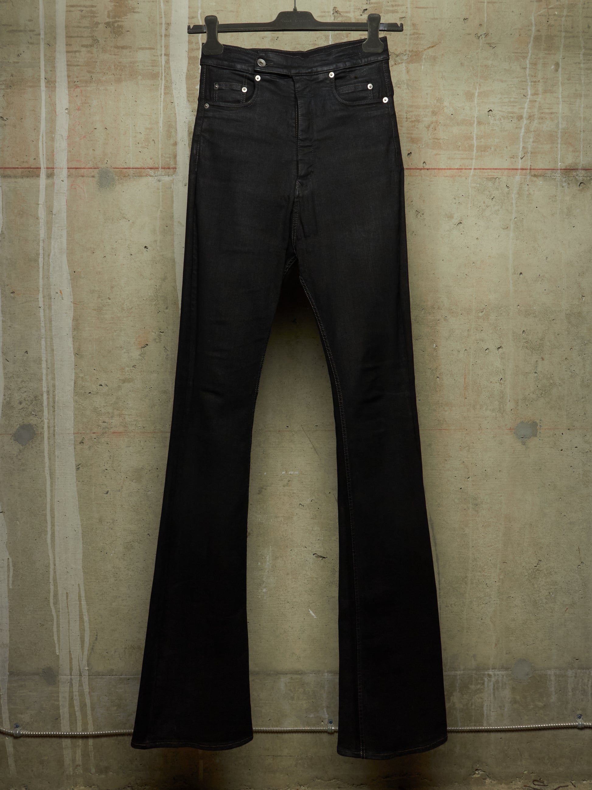 Black flare jeans hanging on a black hanger against a textured concrete wall.