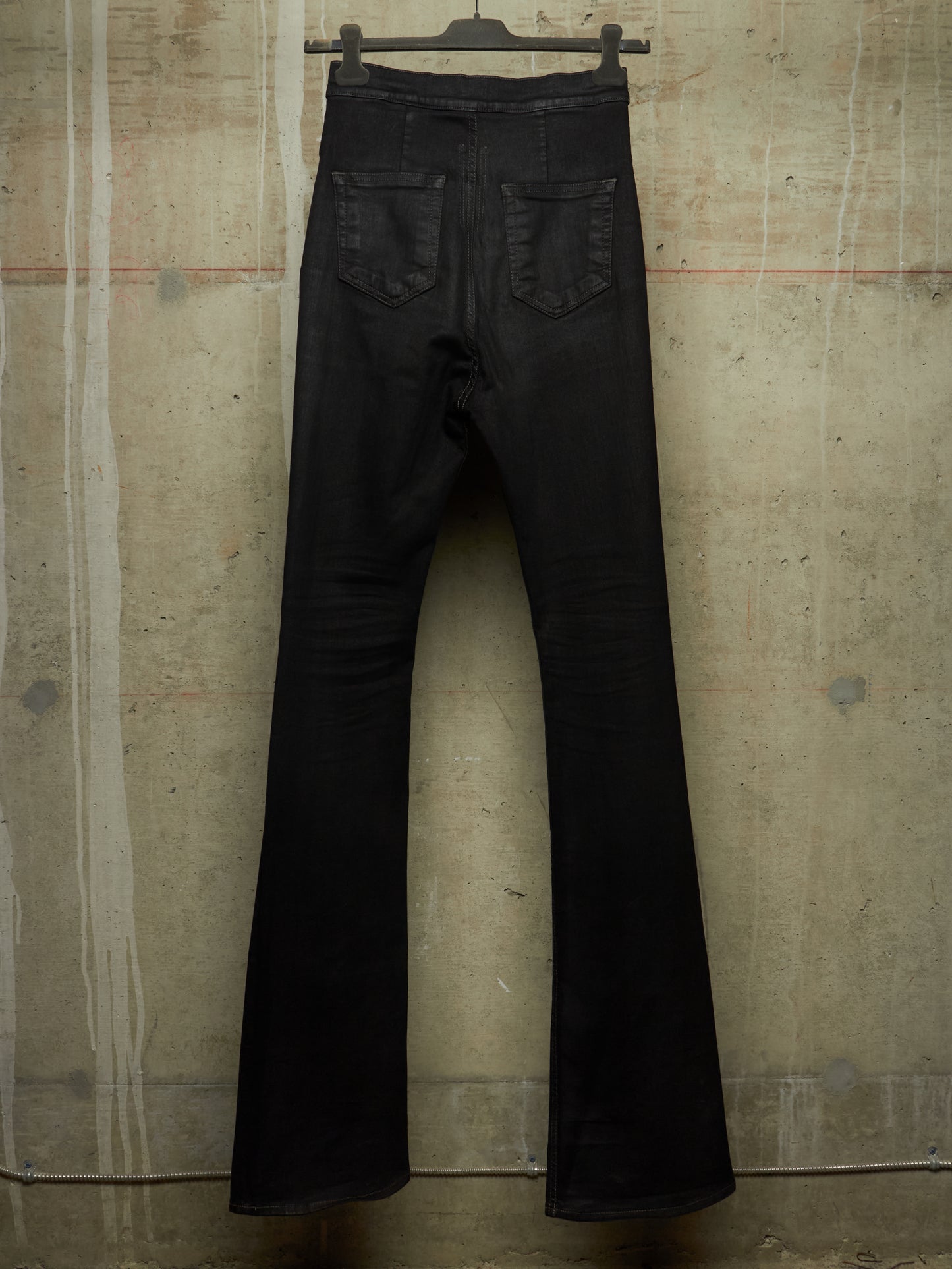 Black flare jeans hanging from a black hanger against a textured concrete wall.