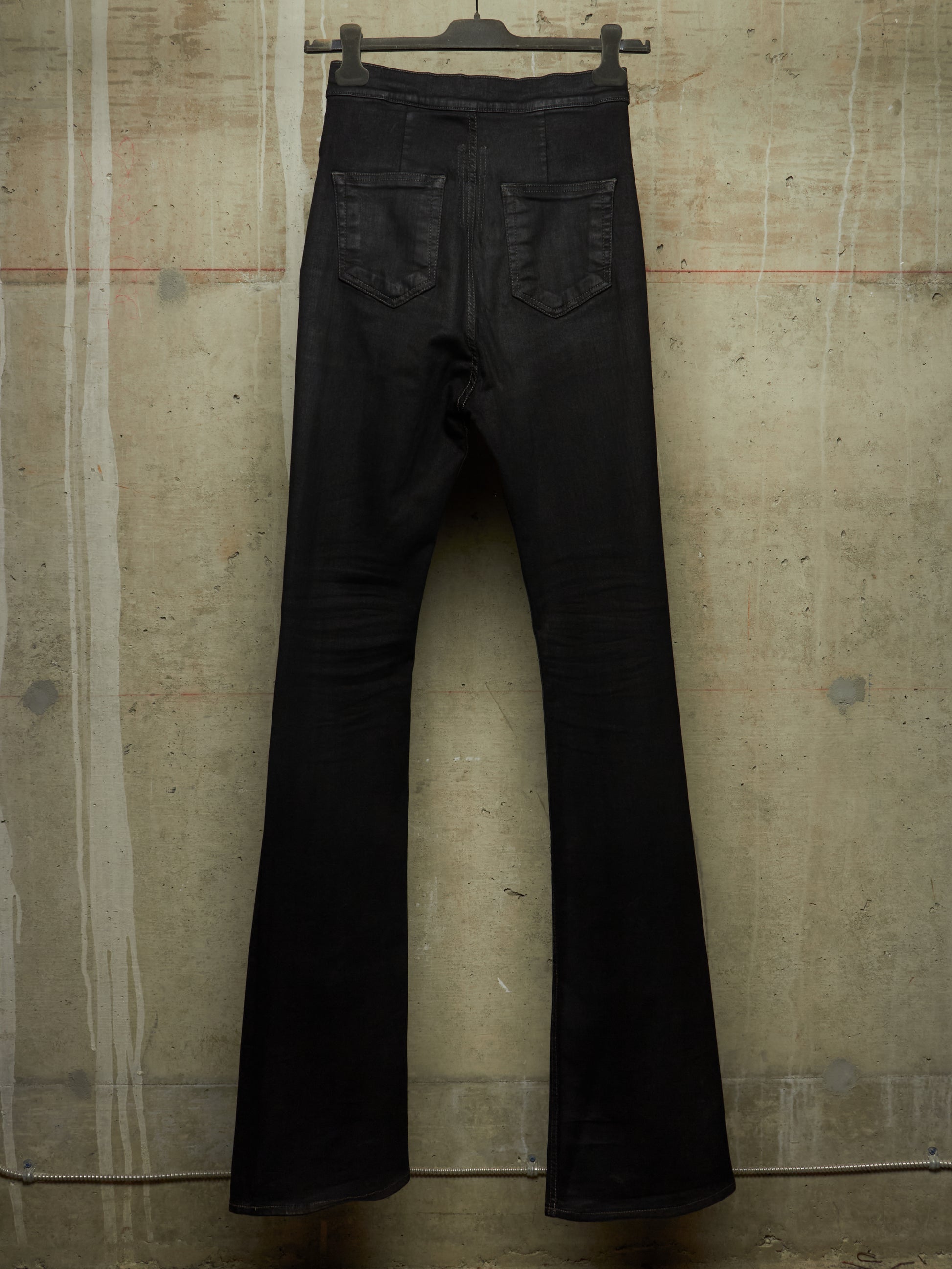 Black flare jeans hanging from a black hanger against a textured concrete wall.