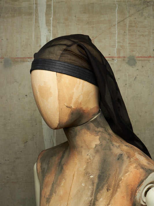 A black sheer headscarf or veil displayed on a painted mannequin torso against a textured concrete wall.