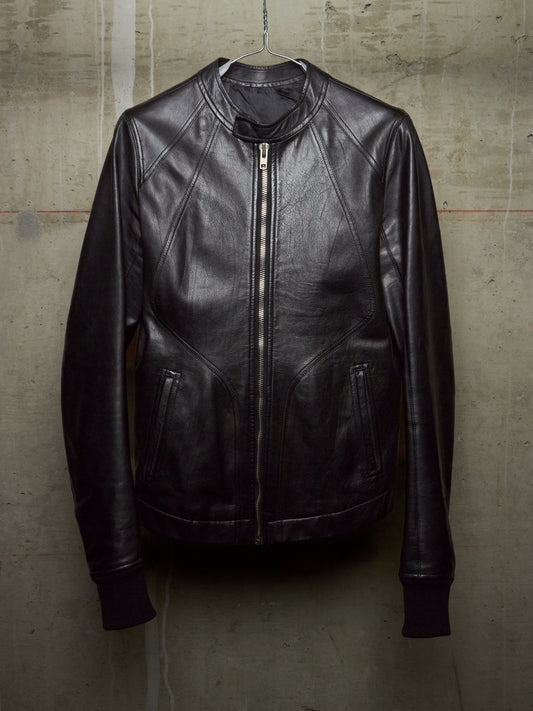 A black leather jacket with a front zipper, ribbed collar, cuffs, and hem, hanging against a textured concrete wall.