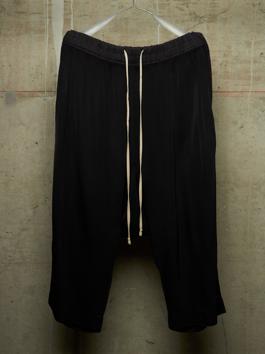Black wide-leg cropped pants with an elastic waistband and cream drawstrings, hanging against a textured concrete wall.