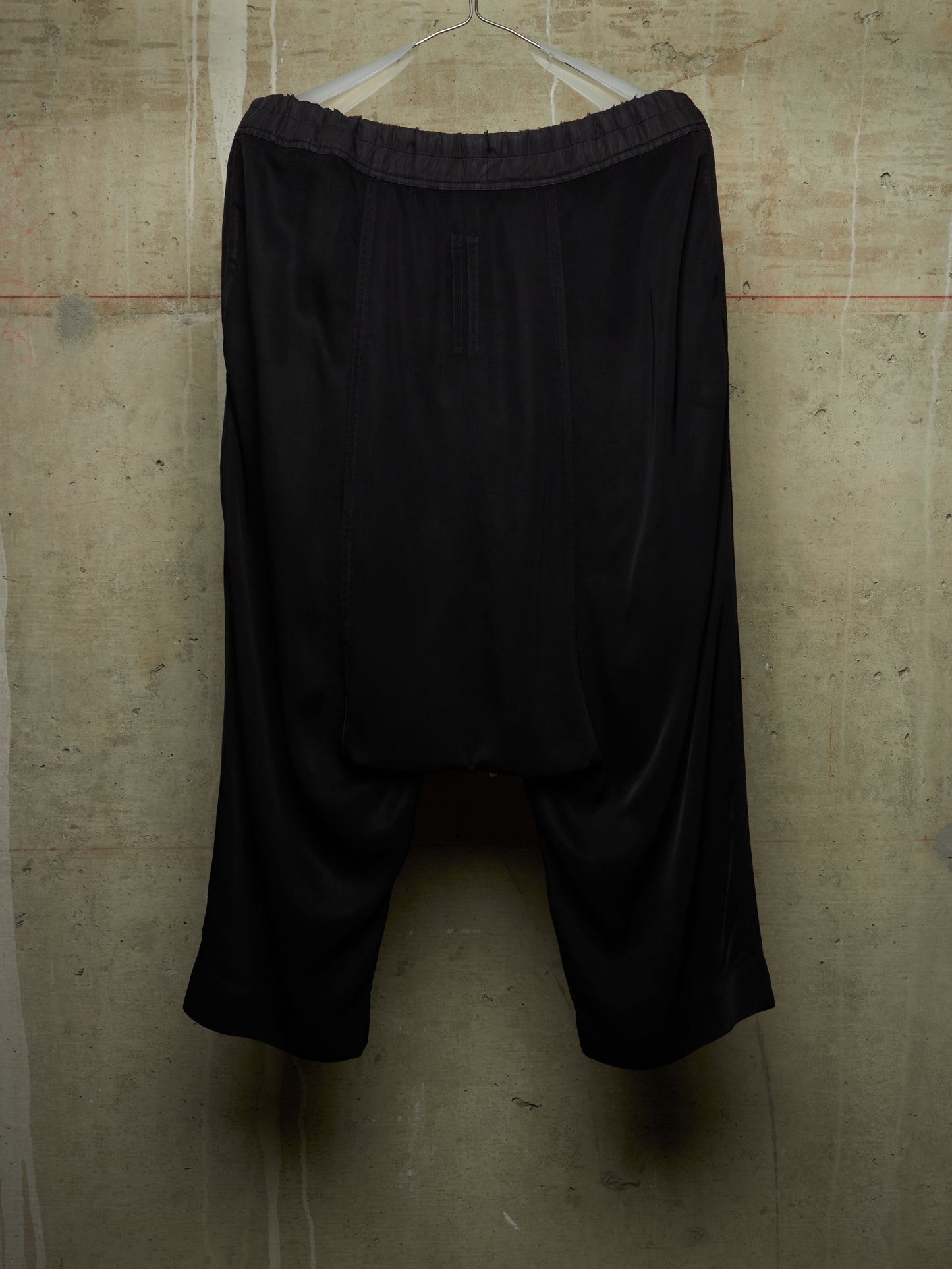 Black harem-style shorts with an elastic waistband, hanging on a metal hanger against a textured concrete wall.