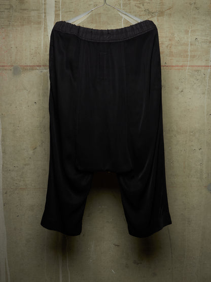 Black harem-style shorts with an elastic waistband, hanging on a metal hanger against a textured concrete wall.