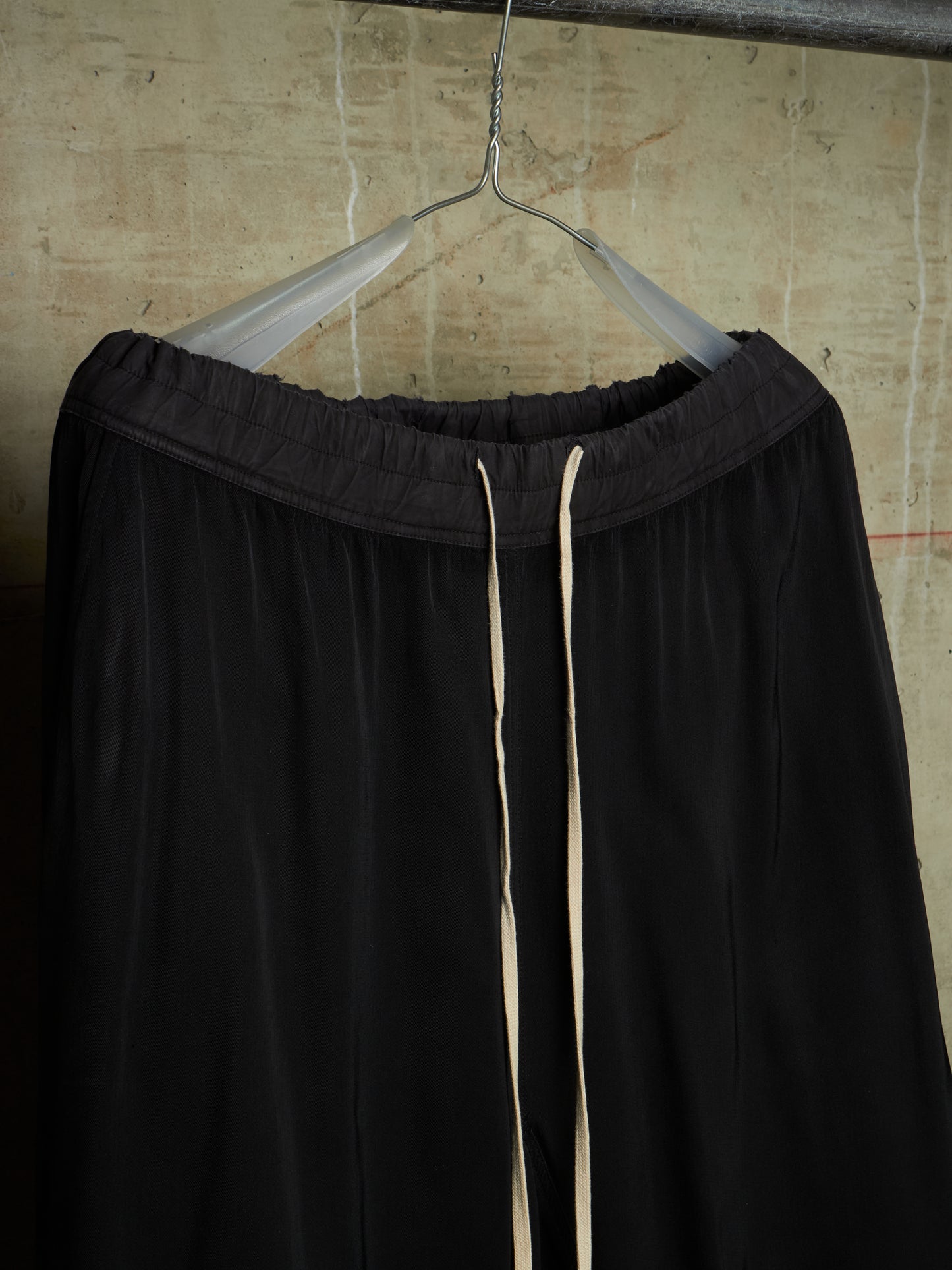 Black wide-leg lounge pants with a drawstring waist, hanging on a clear plastic hanger against a textured concrete wall.