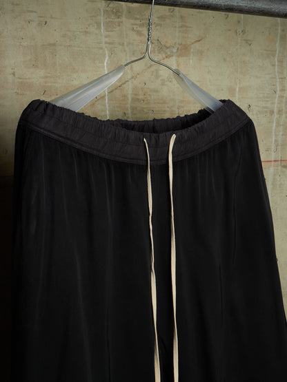 Black wide-leg lounge pants with a drawstring waist, hanging on a clear plastic hanger against a textured concrete wall.