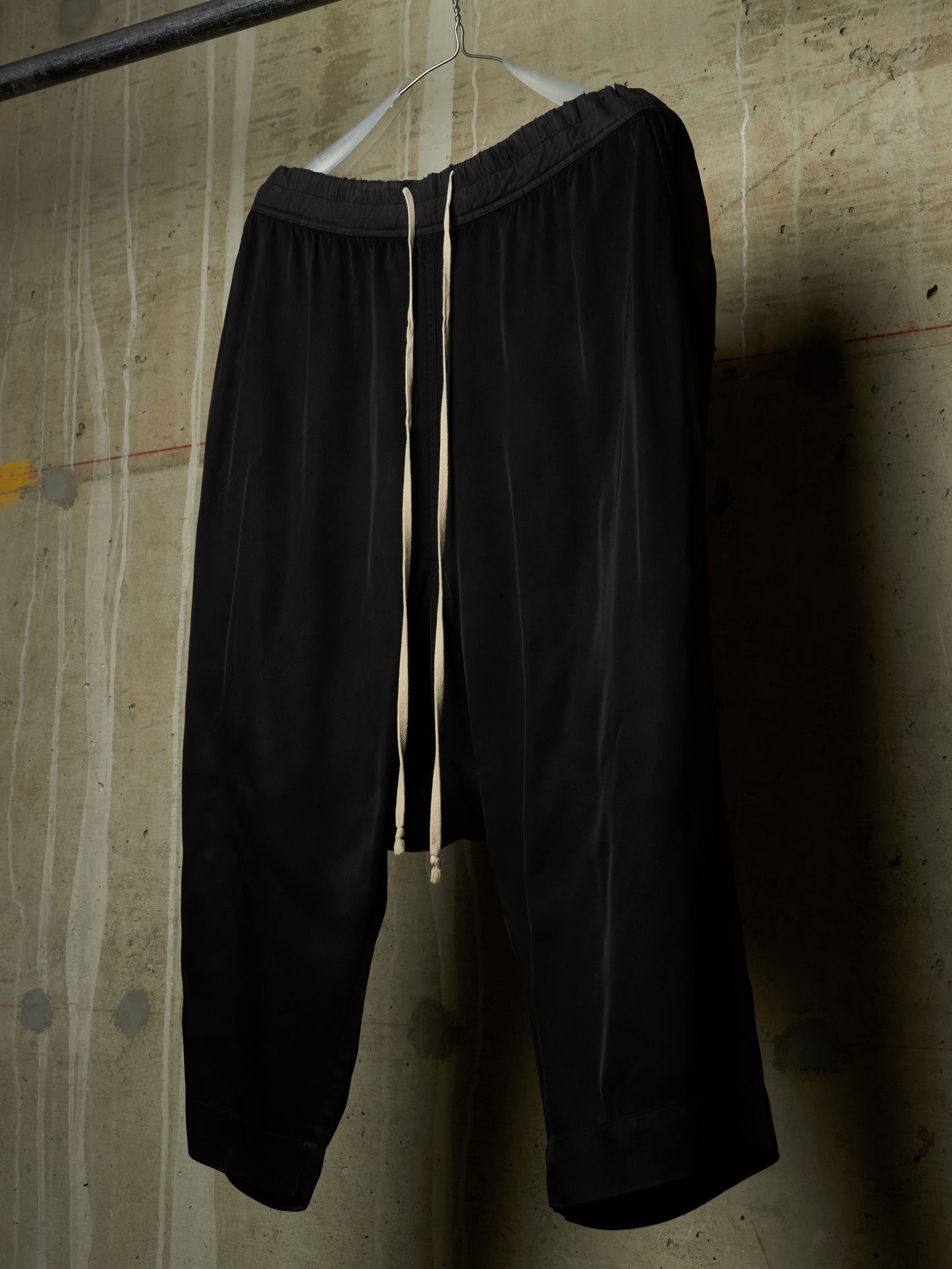 Black wide-leg cropped pants with a cream drawstring, hanging on a metal hanger against a textured concrete wall.