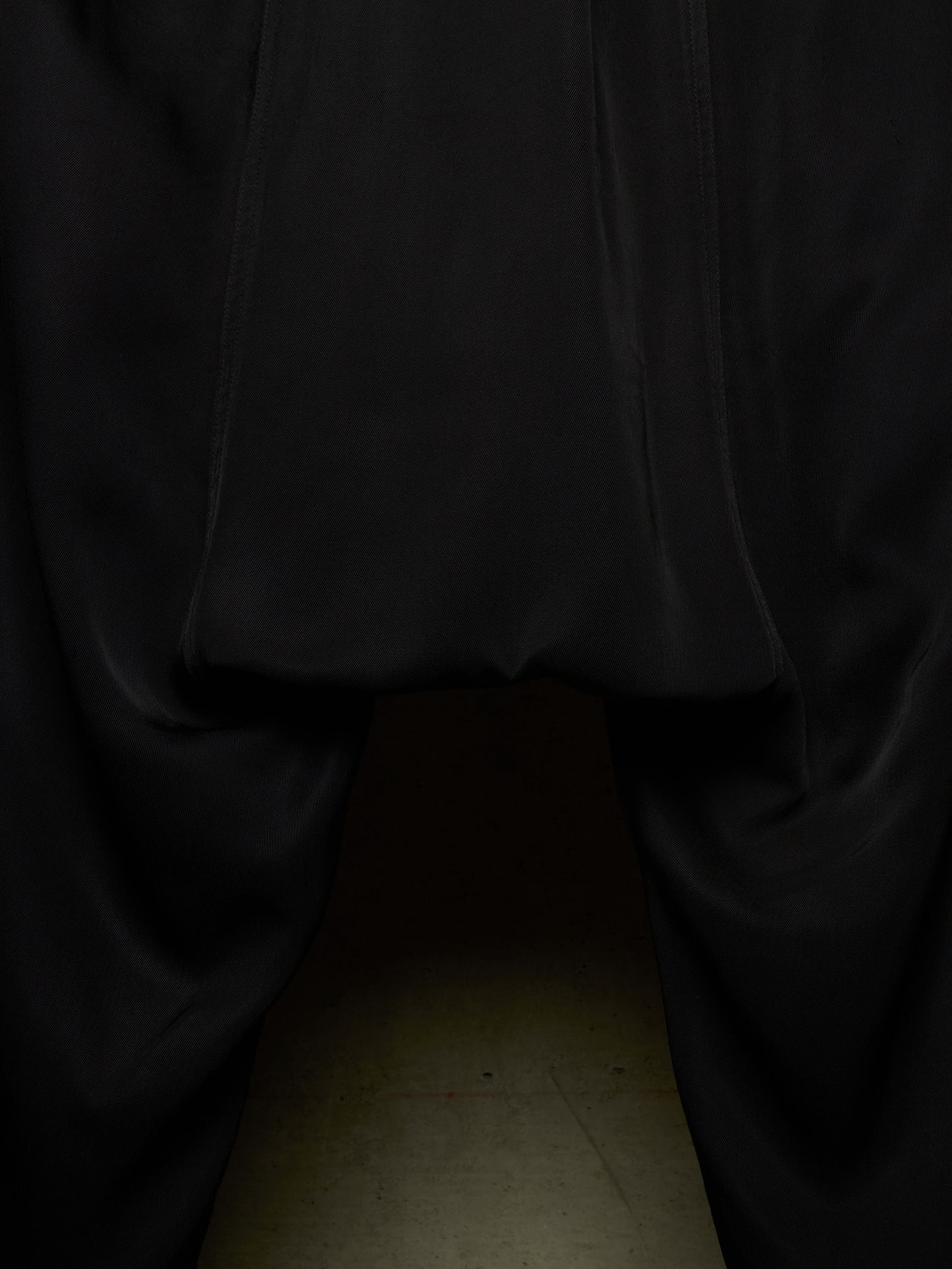 Close-up of a black garment, likely a skirt or draped fabric, gathered at the top with a hollow opening below, photographed against a dark background with a faint beige floor visible at the bottom.