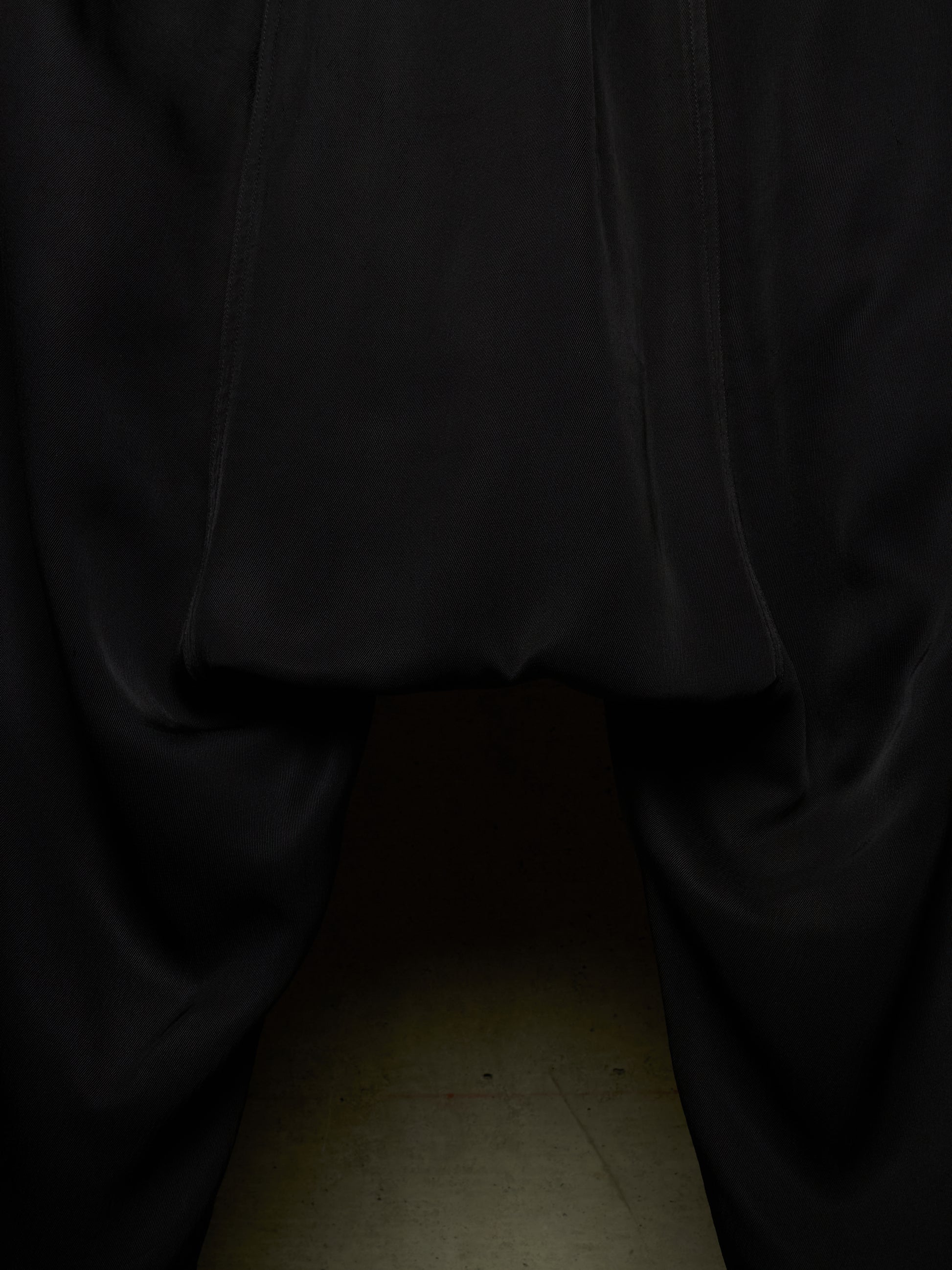 Close-up of a black garment, likely a skirt or draped fabric, gathered at the top with a hollow opening below, photographed against a dark background with a faint beige floor visible at the bottom.