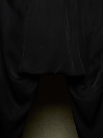 Close-up of a black garment, likely a skirt or draped fabric, gathered at the top with a hollow opening below, photographed against a dark background with a faint beige floor visible at the bottom.