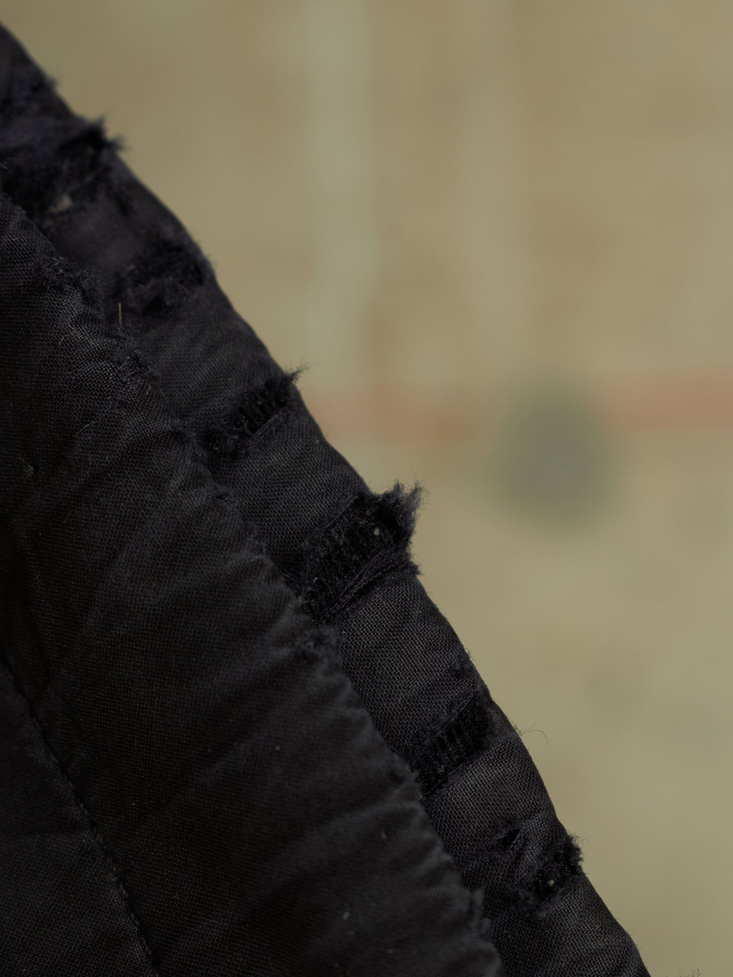 Close-up detail of a black textured fabric item with gathered pleats and frayed edges against a blurred beige background.