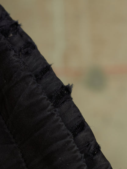 Close-up detail of a black textured fabric item with gathered pleats and frayed edges against a blurred beige background.