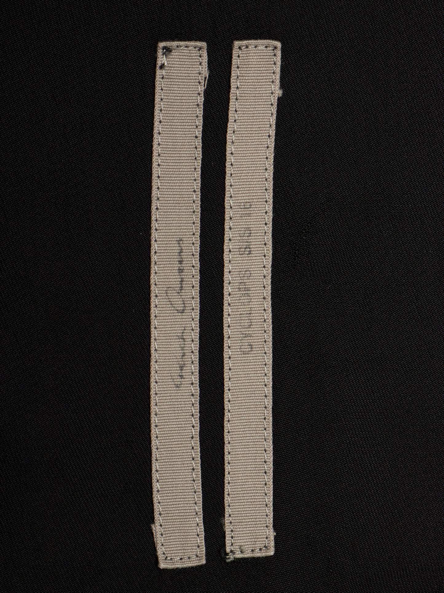 Two narrow beige fabric labels with visible stitching and handwritten markings, placed on a black fabric background.