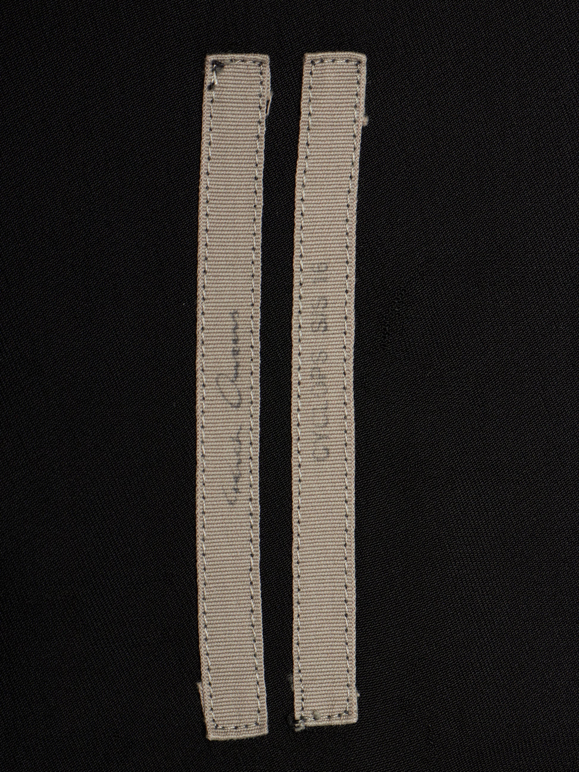 Two narrow beige fabric labels with visible stitching and handwritten markings, placed on a black fabric background.