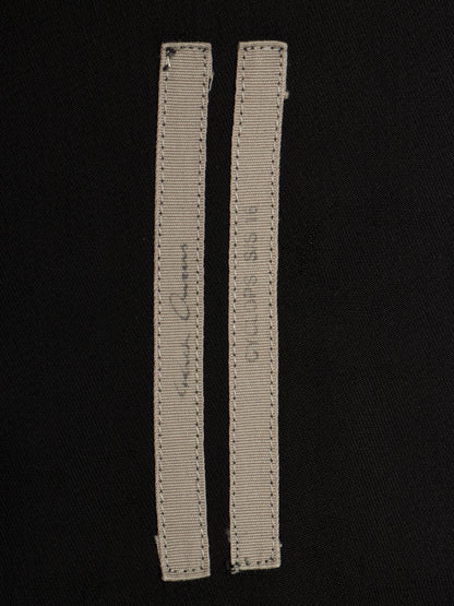 Two narrow beige fabric labels with visible stitching and handwritten markings, placed on a black fabric background.