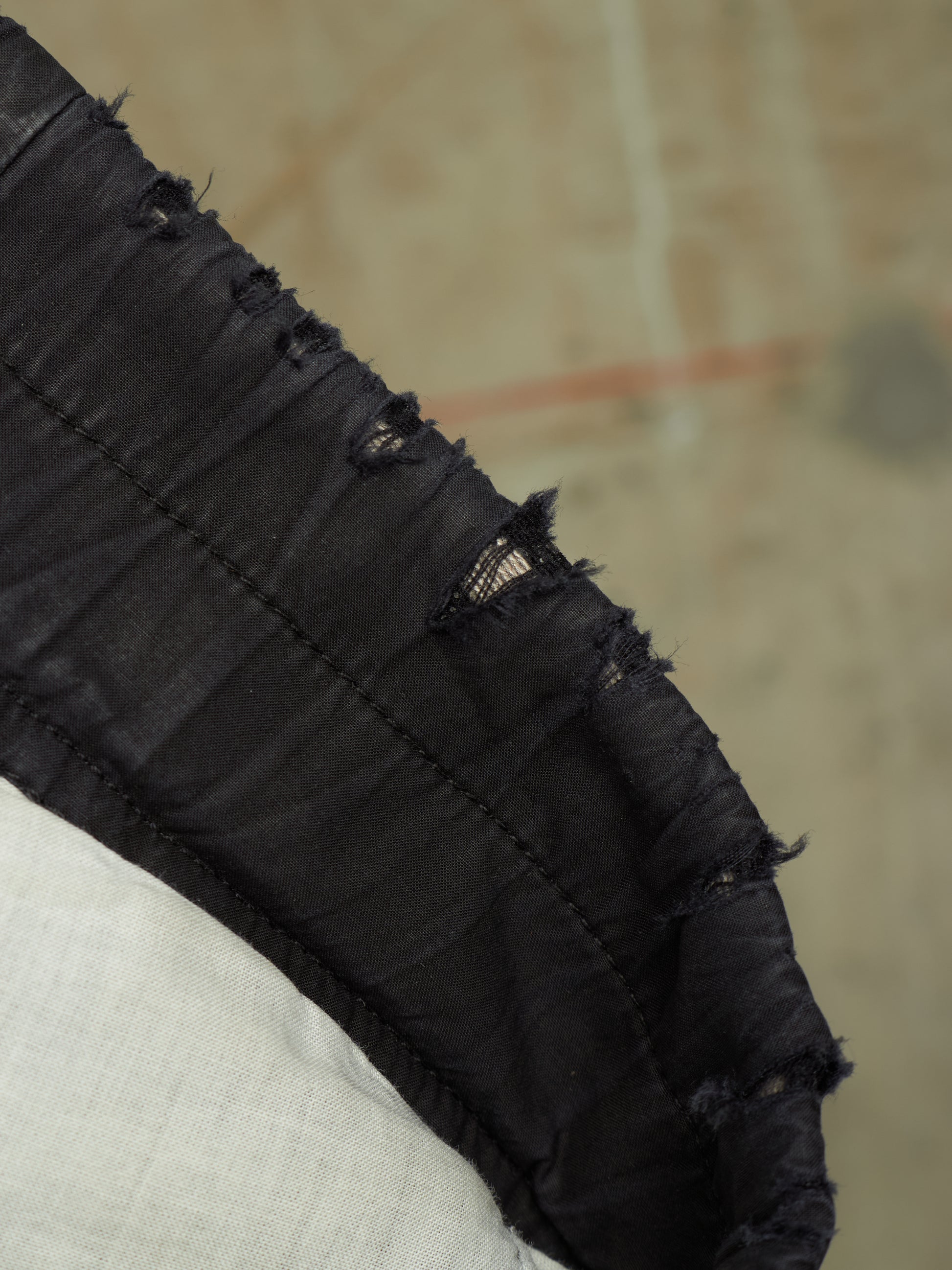 Close-up detail of a black distressed garment, showing gathered fabric, raw frayed edges, and contrast stitching against a neutral beige background.