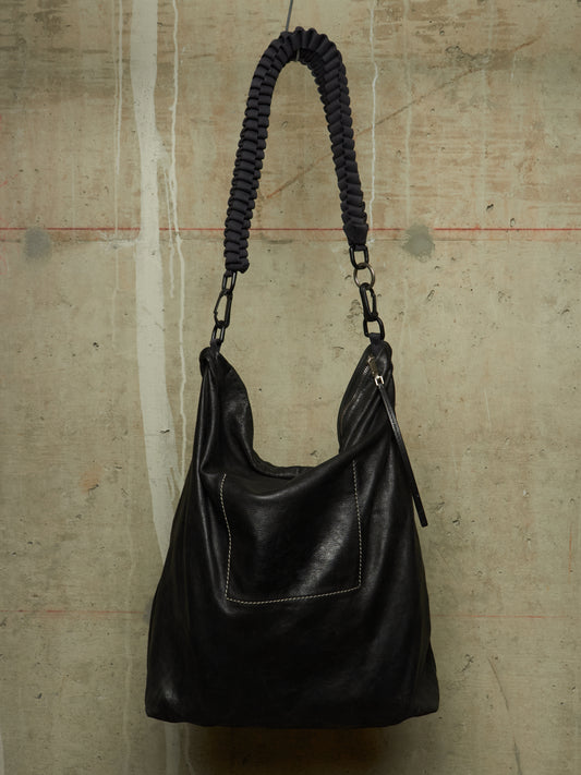 A black leather slouchy shoulder bag with a thick braided black strap and silver-tone hardware, hanging against a textured concrete wall.