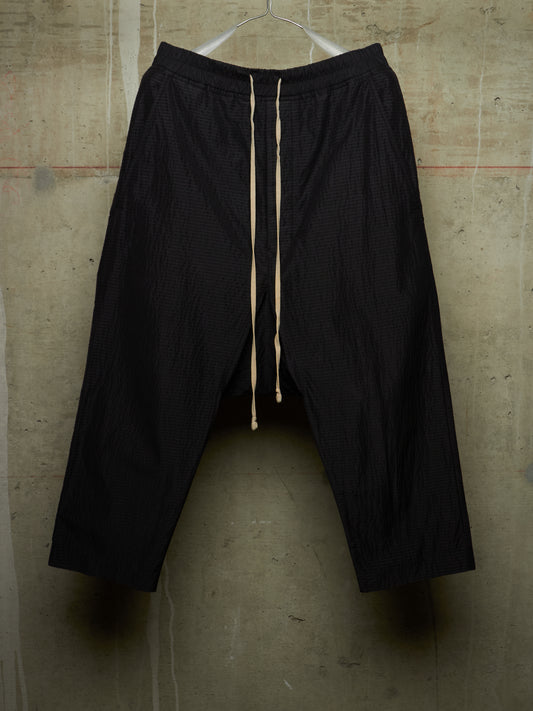 Black textured harem-style pants with a cream drawstring, hanging on a white hanger against a concrete wall.
