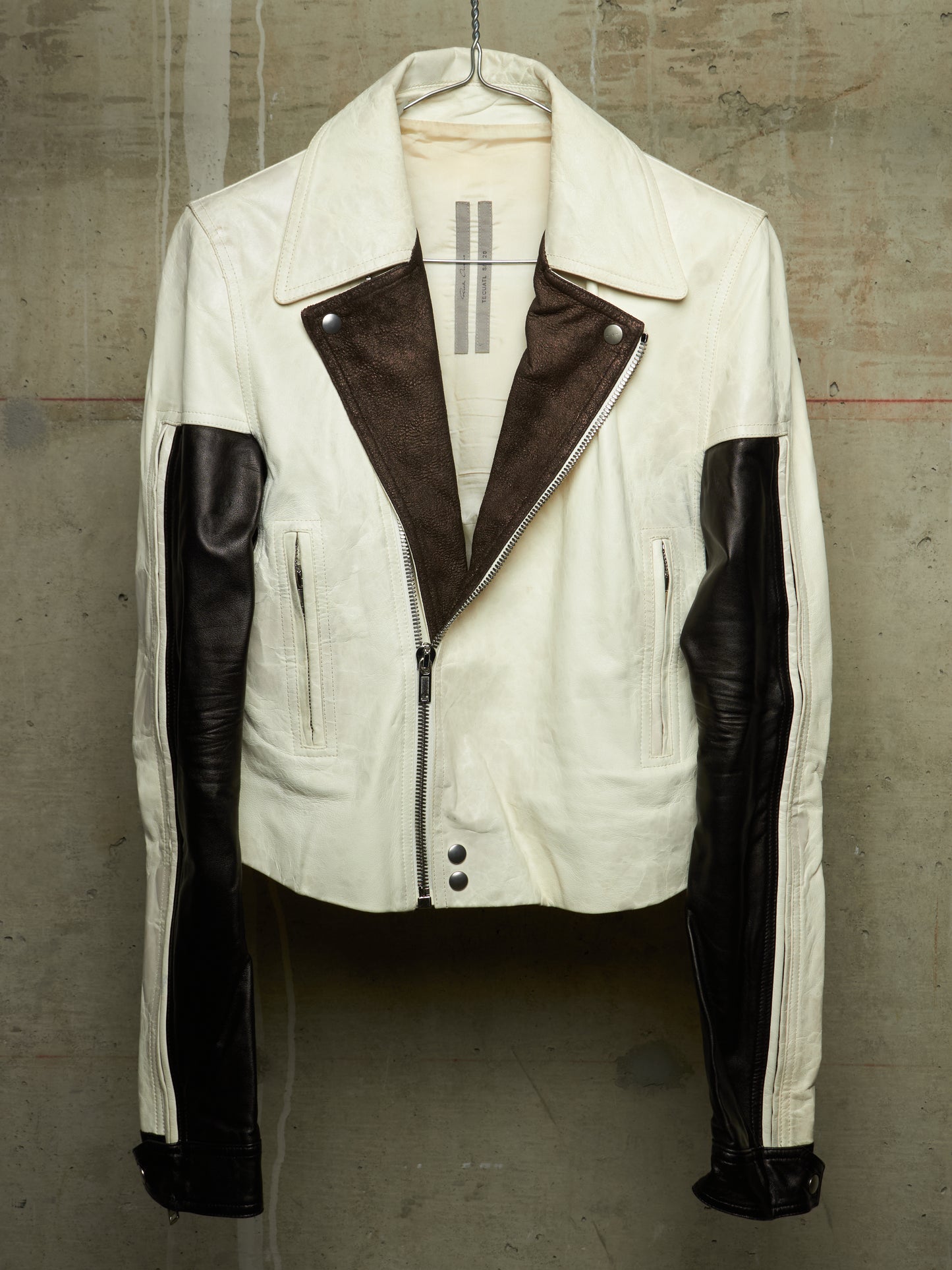 A vintage-style motorcycle jacket in off-white and dark brown leather, hanging on a hanger against a textured concrete wall.