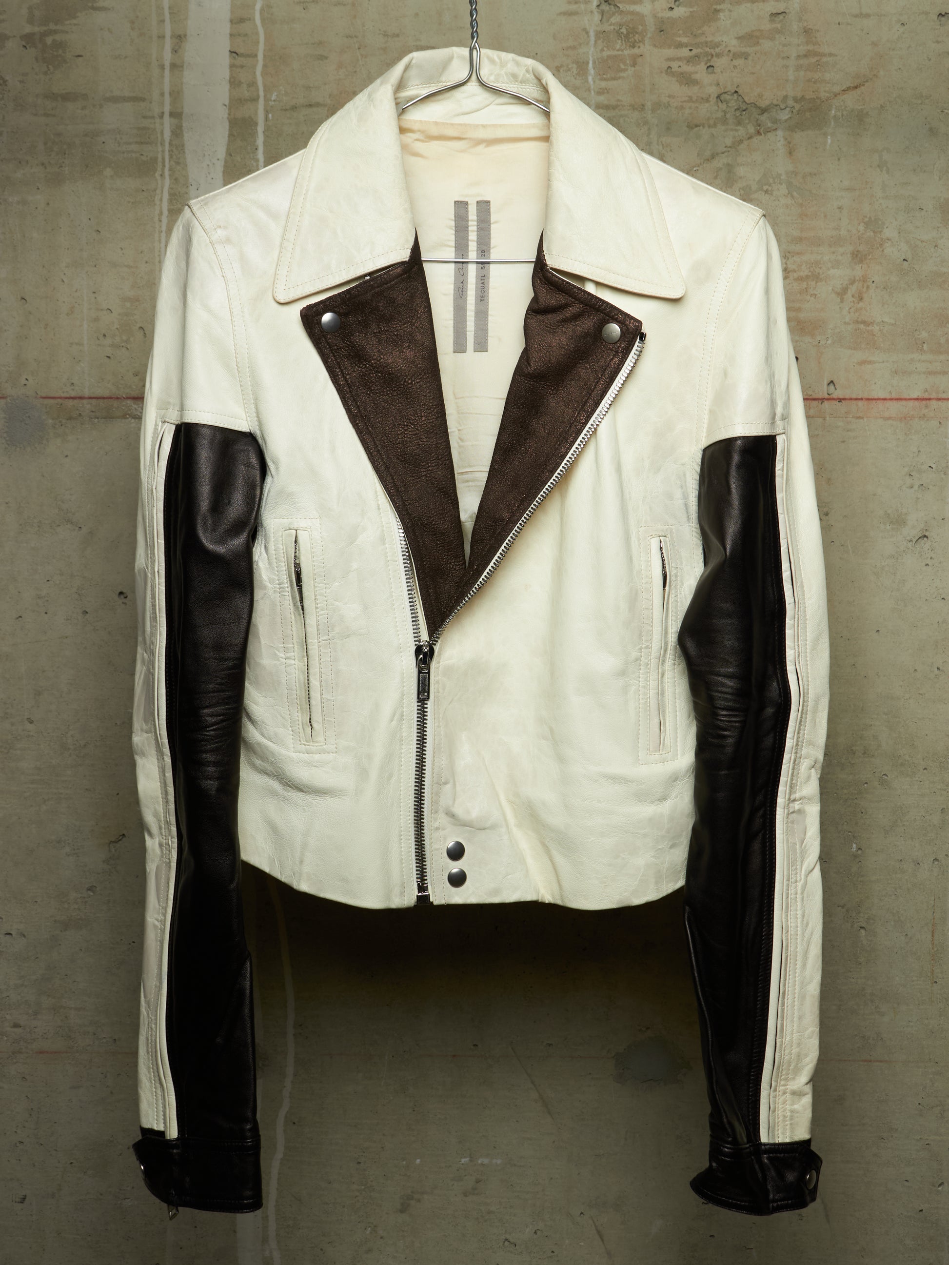 A vintage-style motorcycle jacket in off-white and dark brown leather, hanging on a hanger against a textured concrete wall.