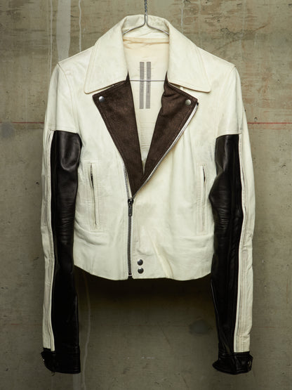 A vintage-style motorcycle jacket in off-white and dark brown leather, hanging on a hanger against a textured concrete wall.