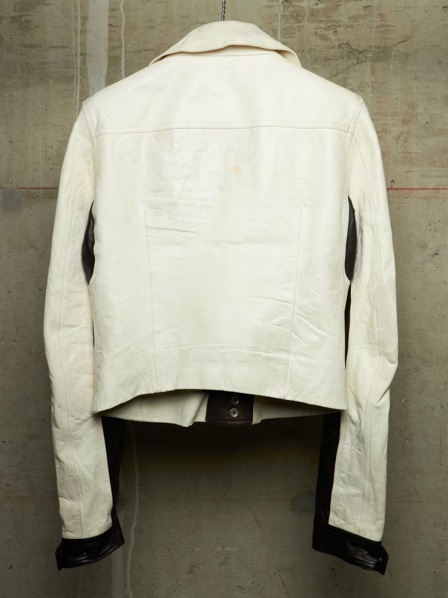The back view of a cream-colored leather jacket with black leather accents on the inner sleeves and cuffs, displayed on a mannequin against a textured concrete wall.