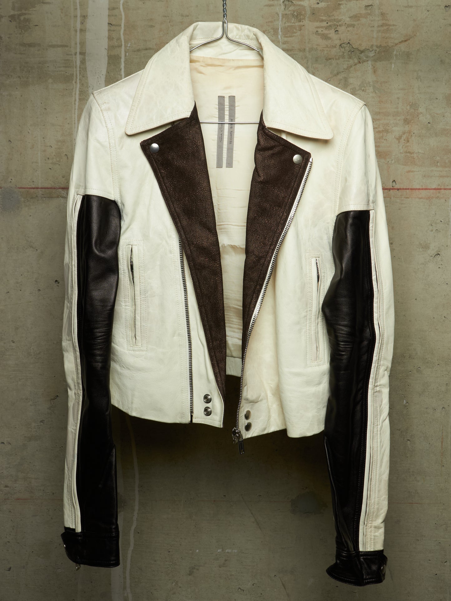 A cream-colored leather jacket with dark brown suede-like panels on the front and black leather sleeves, hanging against a textured concrete wall.