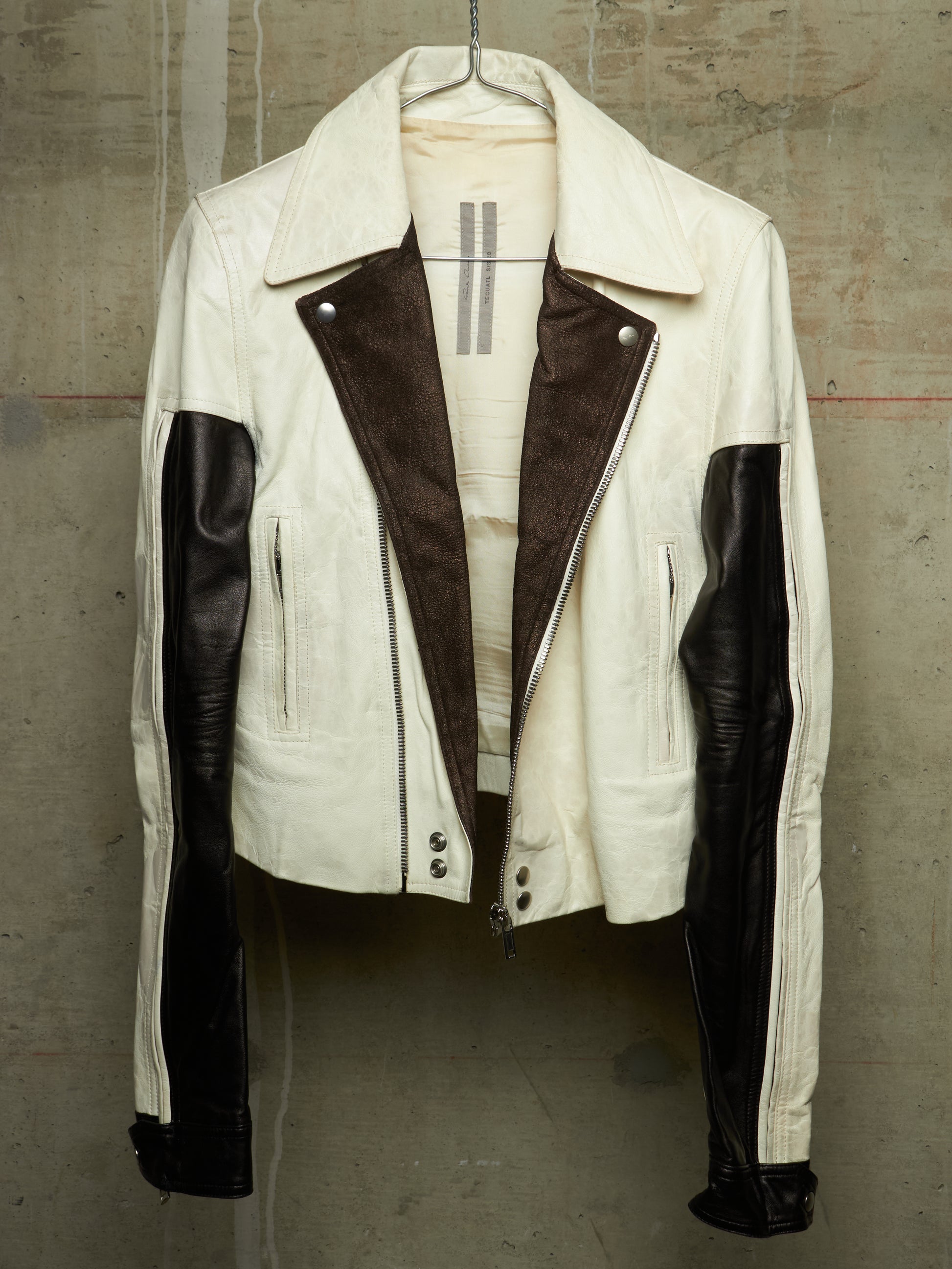 A cream-colored leather jacket with dark brown suede-like panels on the front and black leather sleeves, hanging against a textured concrete wall.