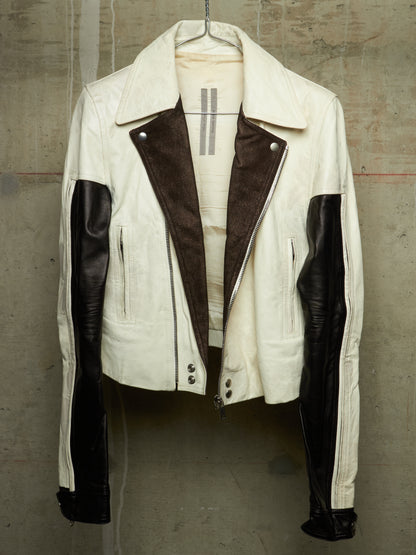A cream-colored leather jacket with dark brown suede-like panels on the front and black leather sleeves, hanging against a textured concrete wall.