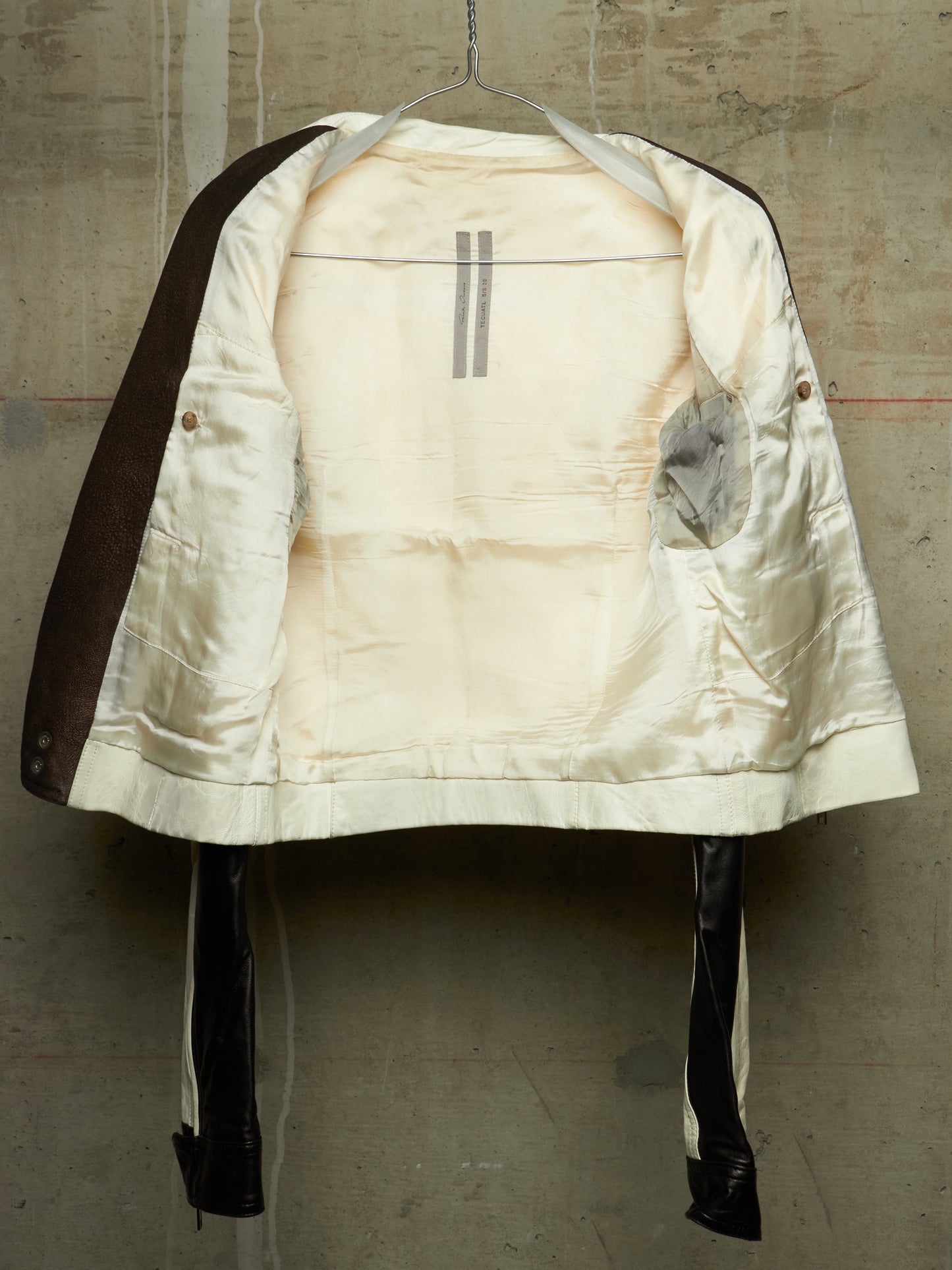 A vintage quilted jacket with a cream satin lining and dark brown sleeves, hanging on a wire hanger against a textured concrete wall.