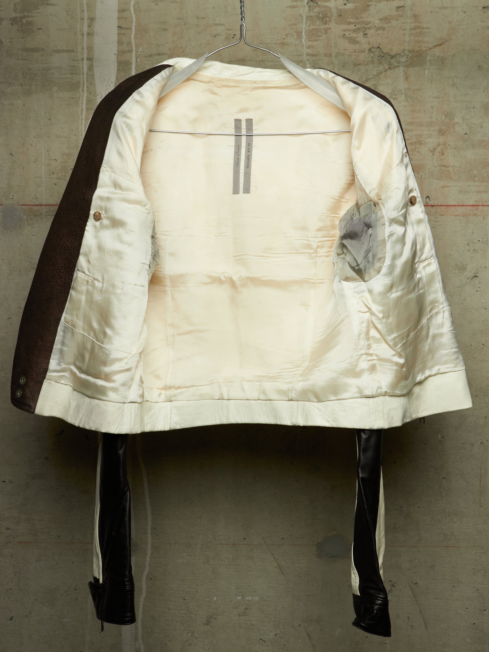 A vintage quilted jacket with a cream satin lining and dark brown sleeves, hanging on a wire hanger against a textured concrete wall.