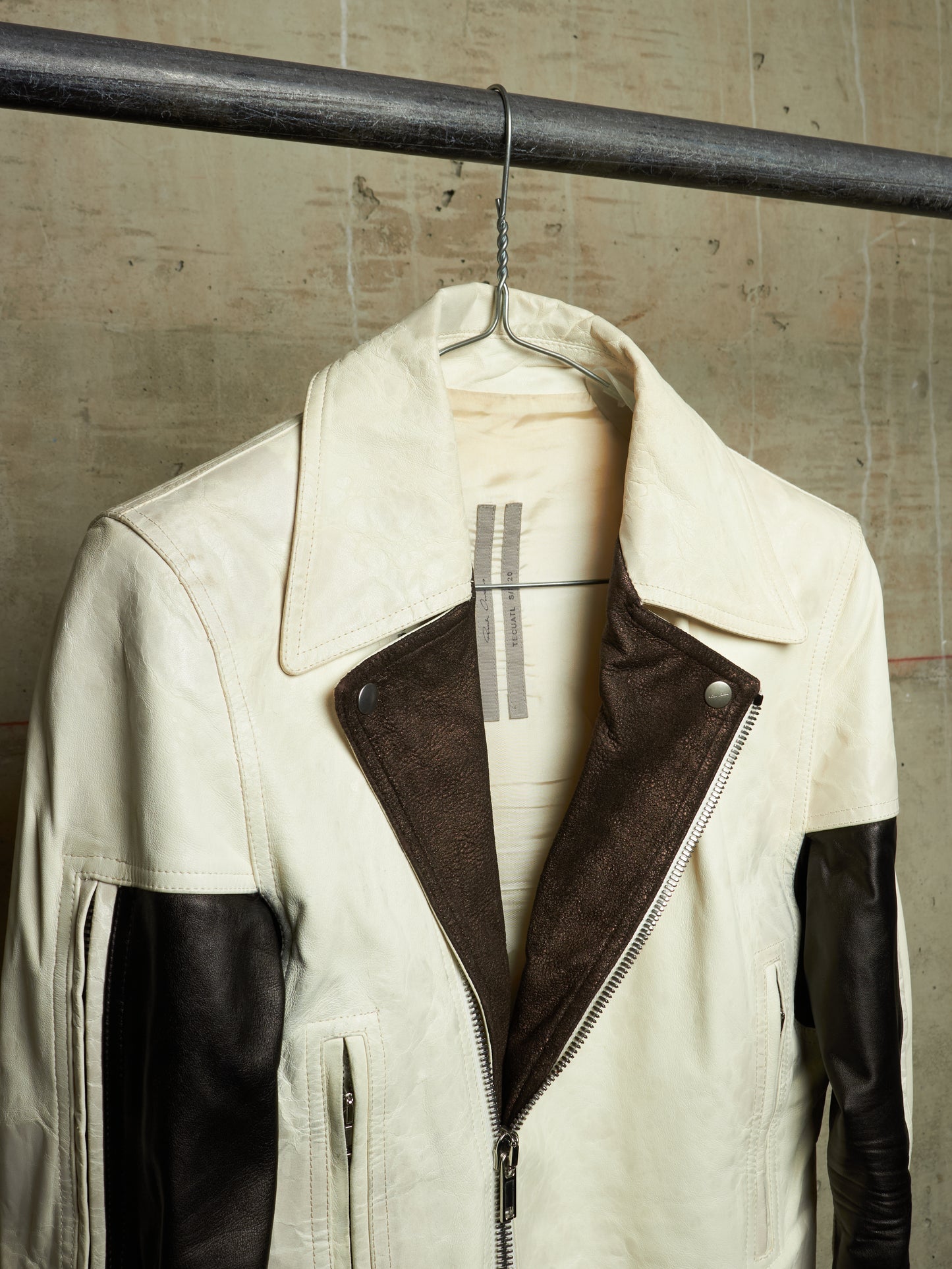 A cream and dark brown color-block leather jacket hanging on a metal hanger from a dark industrial rod, set against a textured concrete wall.