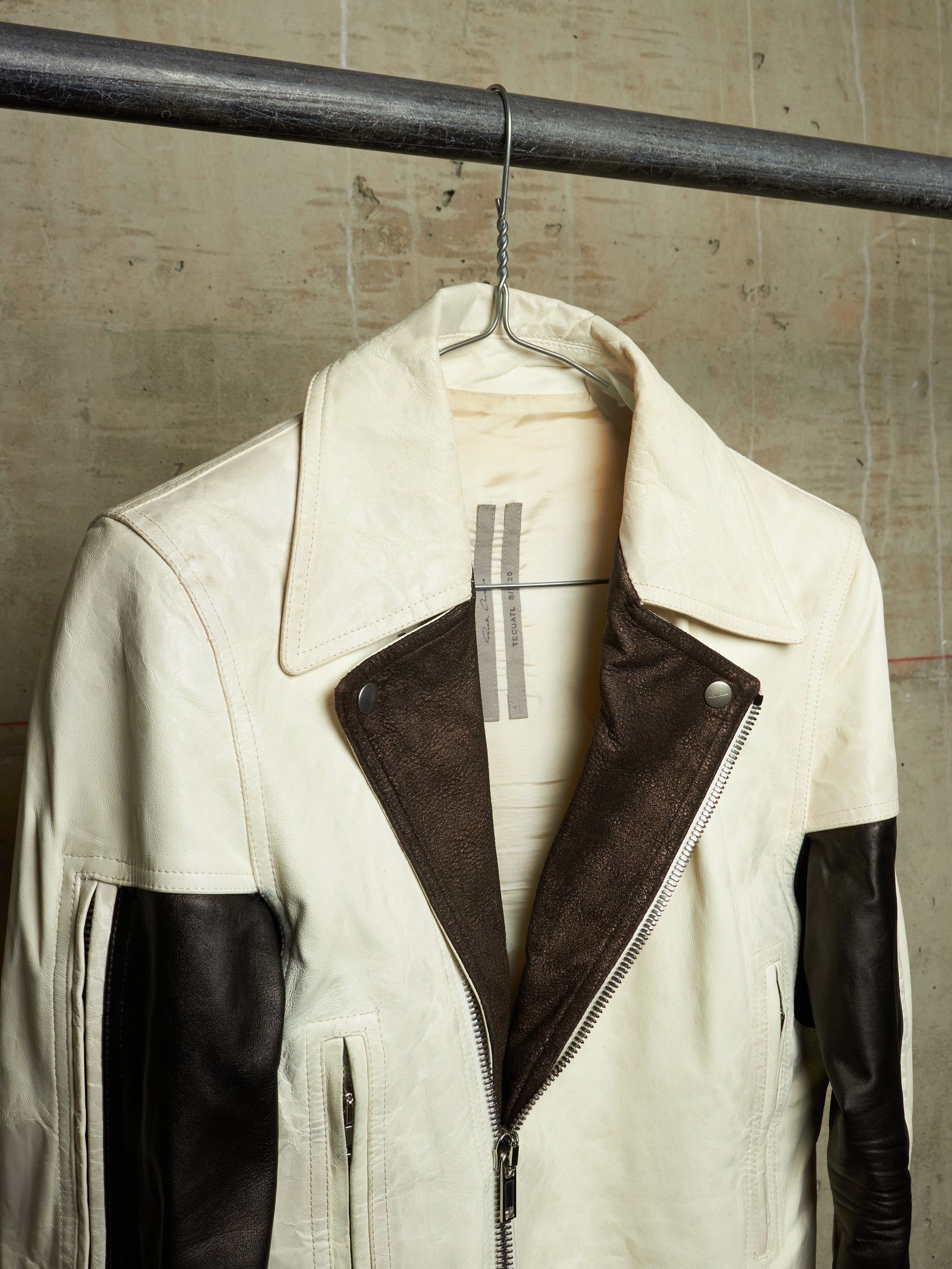 A cream and dark brown color-block leather jacket hanging on a metal hanger from a dark industrial rod, set against a textured concrete wall.