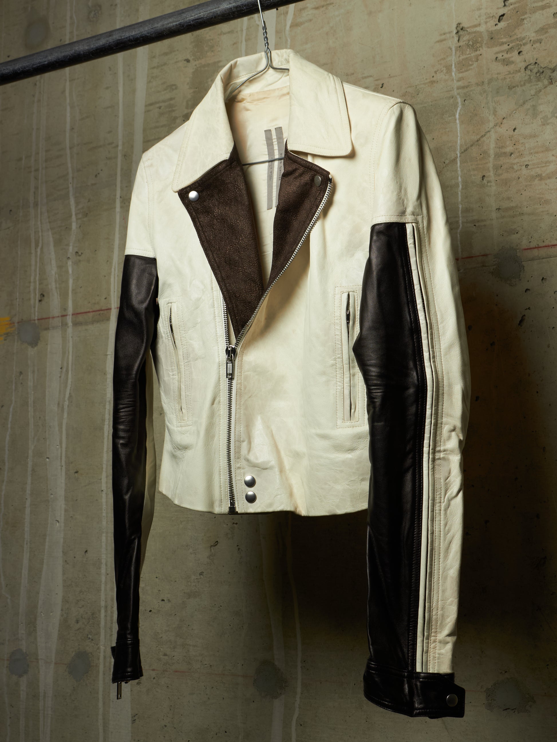 A cream-colored leather jacket with contrasting dark brown suede-like panels on the front and black leather sleeves, hanging on a hanger against a textured concrete wall.