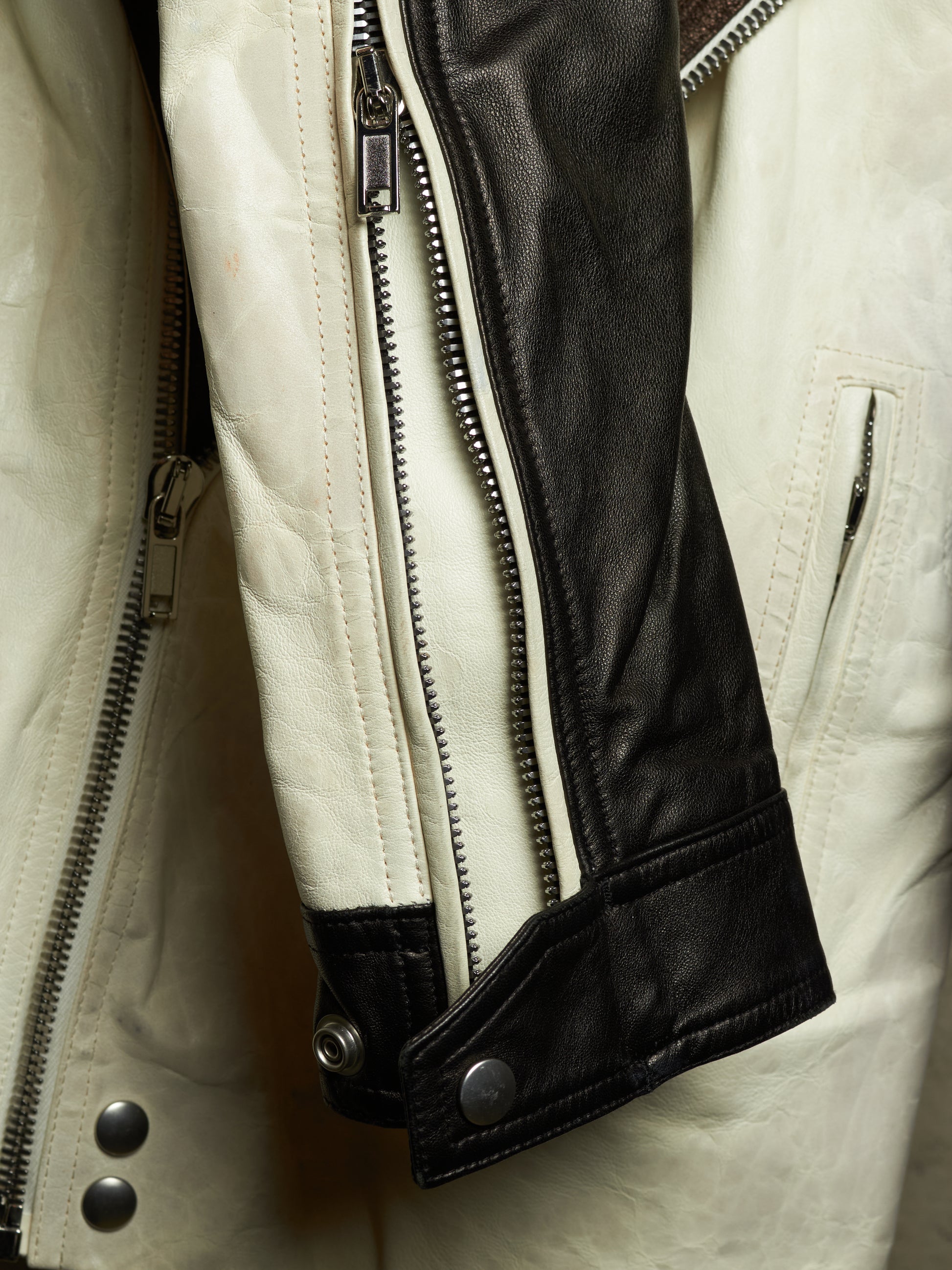 Close-up of a leather jacket showing a cream-colored body, a black leather sleeve panel, silver metal zippers, and snap-button cuffs.