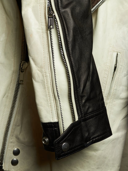 Close-up of a leather jacket showing a cream-colored body, a black leather sleeve panel, silver metal zippers, and snap-button cuffs.